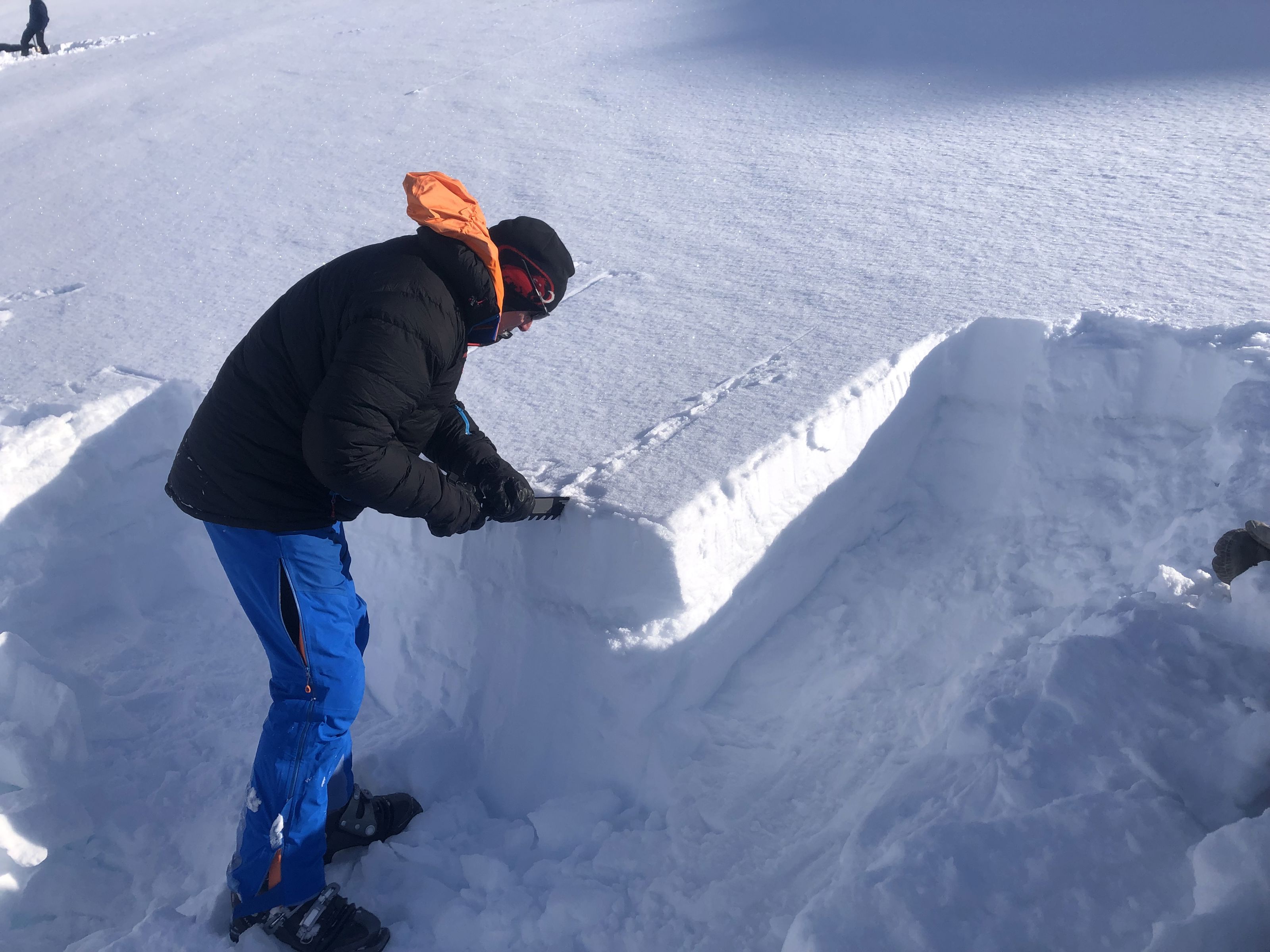 En mann står med ryggen til kamera vendt mot en snøskavel. Det kan se ut som han forsøker å skjære i snøen med et verktøy man helt klart skimter.